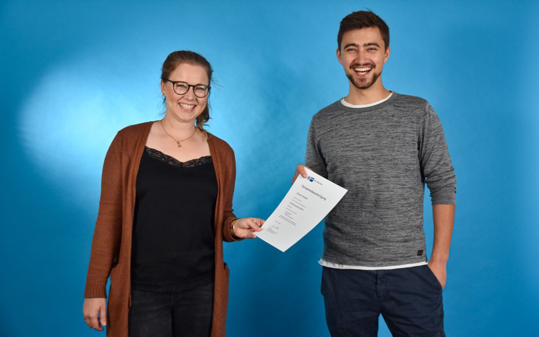 Junger Mann und junge Frau stehen vor einer blauen Wand. Sie lachen und halten gemeinsam ein Zertifikat in der Hand.