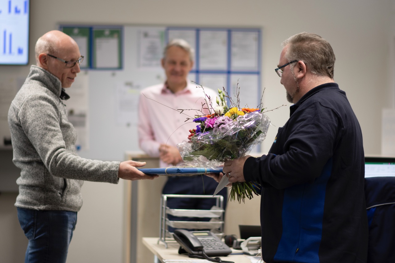 Drei Männer stehen in einem Büro. Ein Mann erhält einen Blumenstrauß.